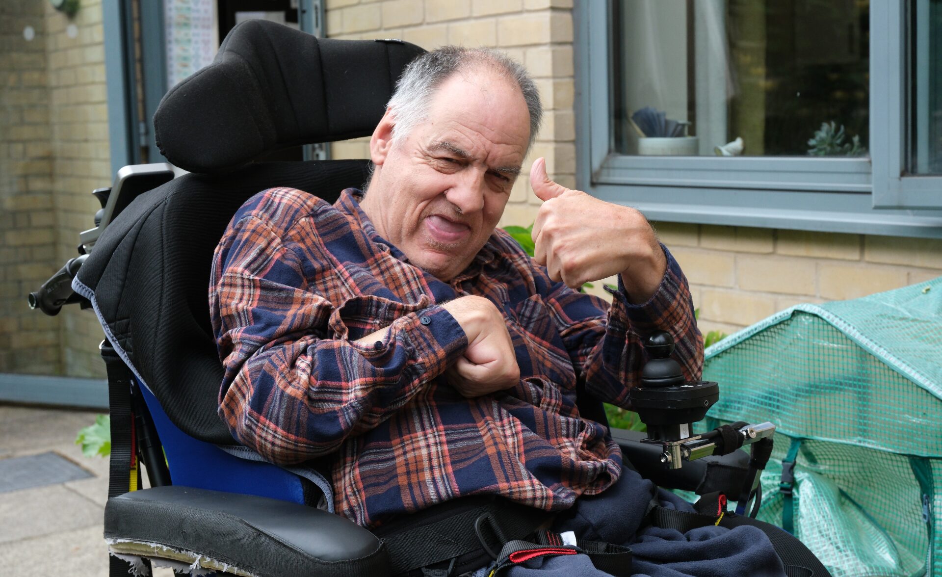 A middle-aged man with grey hair, wearing a red and navy checked shirt, sits in a powered wheelchair outside a supported living residence. He is smiling warmly and giving a thumbs-up with his right hand, expressing confidence and positivity. The setting includes a brick wall, large windows, and a green mini greenhouse, suggesting a homely and supportive environment. The man is a client of Southdown’s learning disability service