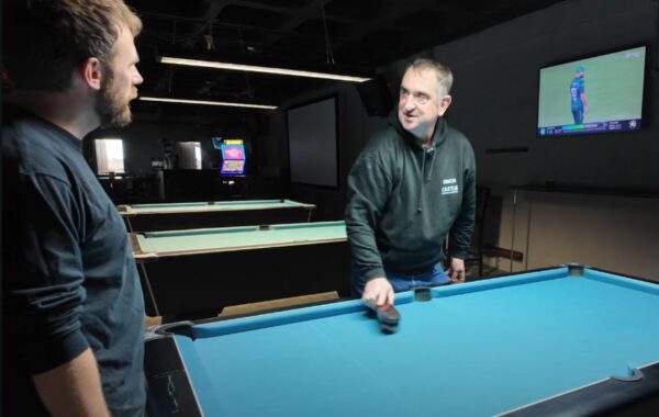 Simon, a Southdown learning disability services client, is smiling while brushing a pool table at his local snooker club, wearing a dark hoodie with his name embroidered on it. A staff member stands nearby, watching and chatting with him. Several pool tables and a TV screen are visible in the background.