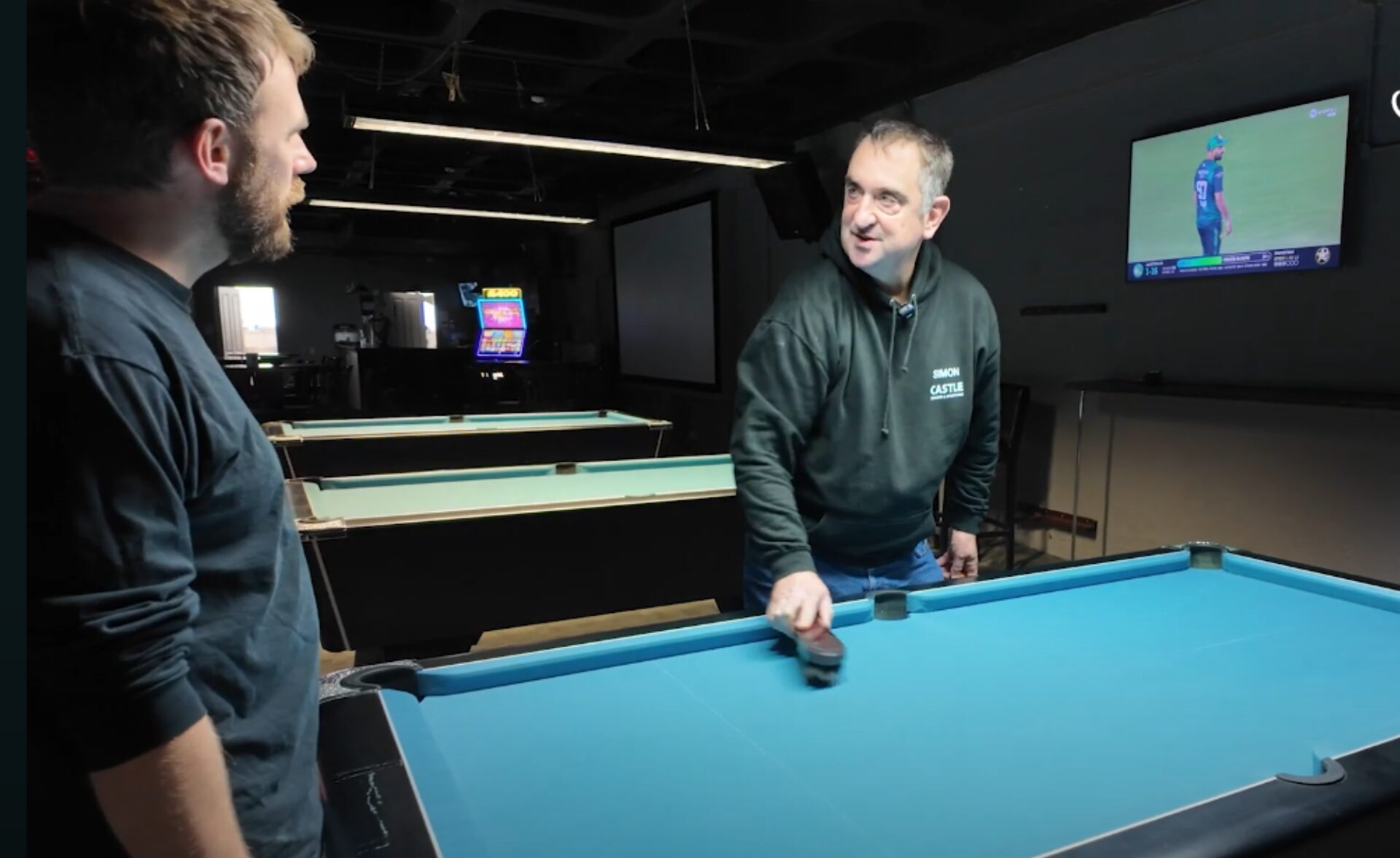 Simon, a Southdown learning disability services client, is smiling while brushing a pool table at his local snooker club, wearing a dark hoodie with his name embroidered on it. A staff member stands nearby, watching and chatting with him. Several pool tables and a TV screen are visible in the background.