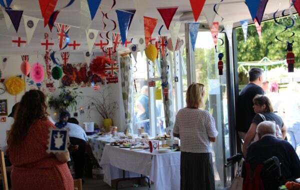 People gathered indoors and outdoors at Bradbury Court’s annual garden party, with colourful bunting, Union Jack flags, and handmade decorations hanging from the ceiling. Tables are set up with cakes, crafts, and refreshments. The atmosphere is lively and welcoming, with clients, staff, and family members chatting and enjoying the event together.