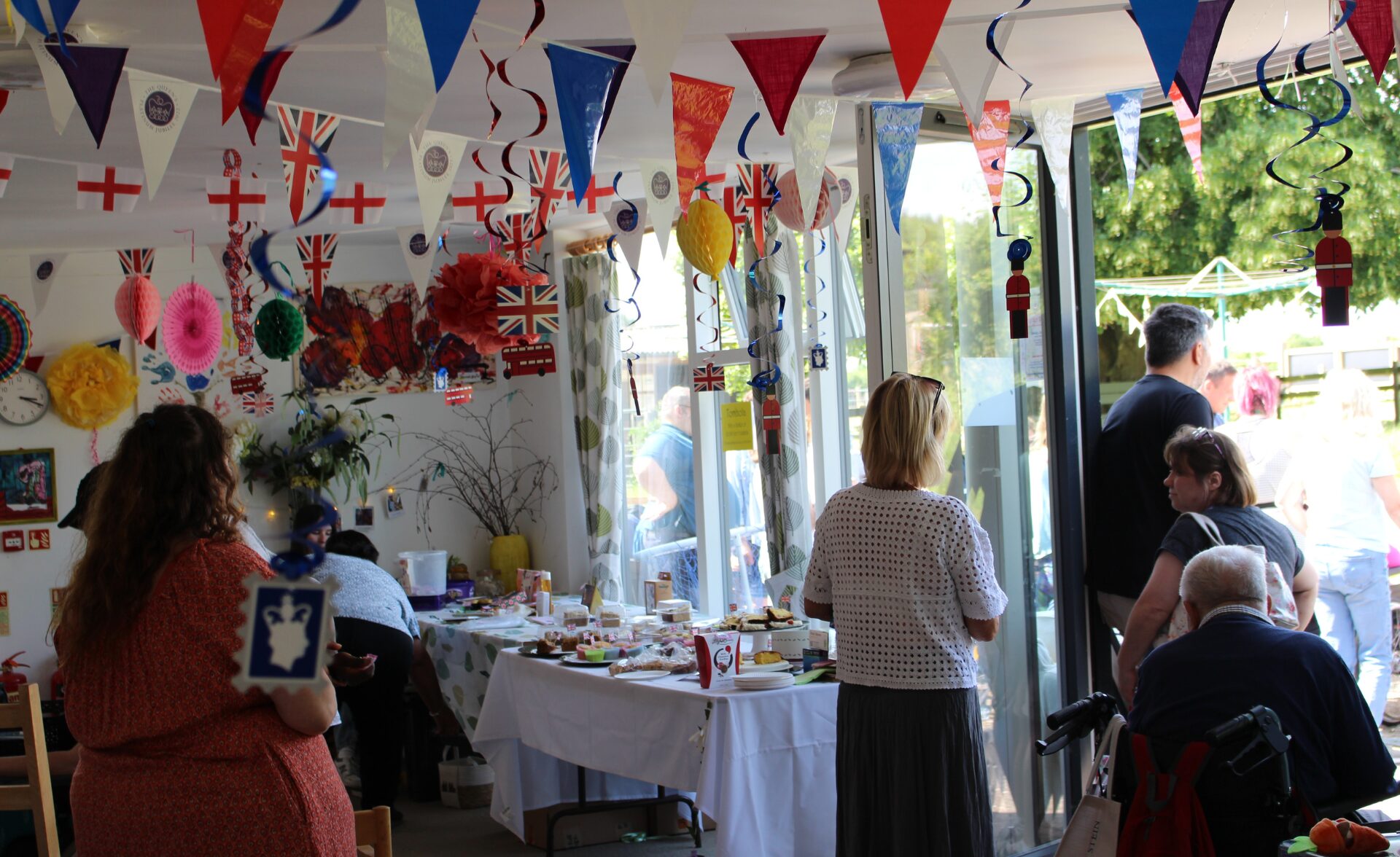 People gathered indoors and outdoors at Bradbury Court’s annual garden party, with colourful bunting, Union Jack flags, and handmade decorations hanging from the ceiling. Tables are set up with cakes, crafts, and refreshments. The atmosphere is lively and welcoming, with clients, staff, and family members chatting and enjoying the event together.