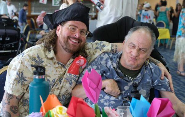 A smiling support worker in a pirate hat poses with a client at the LD Summer Fiesta 2025. Both are wearing tropical shirts and sitting at a decorated table filled with colourful origami flowers. The client, seated in a wheelchair, is playfully pulling a face, and both appear relaxed and happy. In the background, other guests and staff in bright outfits move around a busy, festive hall.