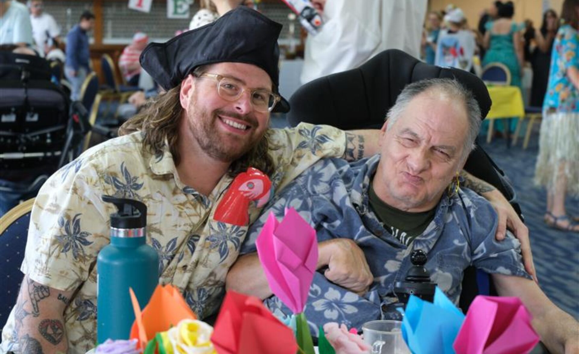 A smiling support worker in a pirate hat poses with a client at the LD Summer Fiesta 2025. Both are wearing tropical shirts and sitting at a decorated table filled with colourful origami flowers. The client, seated in a wheelchair, is playfully pulling a face, and both appear relaxed and happy. In the background, other guests and staff in bright outfits move around a busy, festive hall.