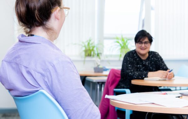 A Connect to Work employment specialist at Southdown supports a client in a relaxed one-to-one meeting. The two are seated at a round table with notes and paperwork, in a bright, welcoming space with natural light and plants in the background.