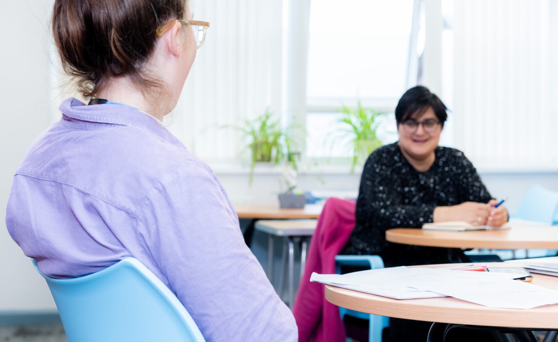 A Connect to Work employment specialist at Southdown supports a client in a relaxed one-to-one meeting. The two are seated at a round table with notes and paperwork, in a bright, welcoming space with natural light and plants in the background.