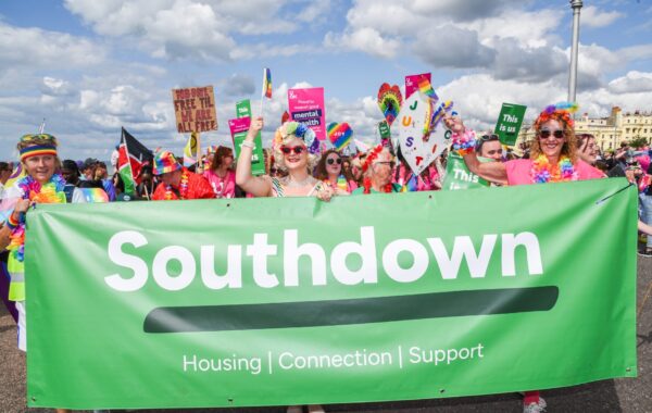Southdown team marching in Brighton Pride Parade, holding a green Southdown banner with the words ‘Housing | Connection | Support’, surrounded by colourful rainbow outfits, flags, and inclusion signs.