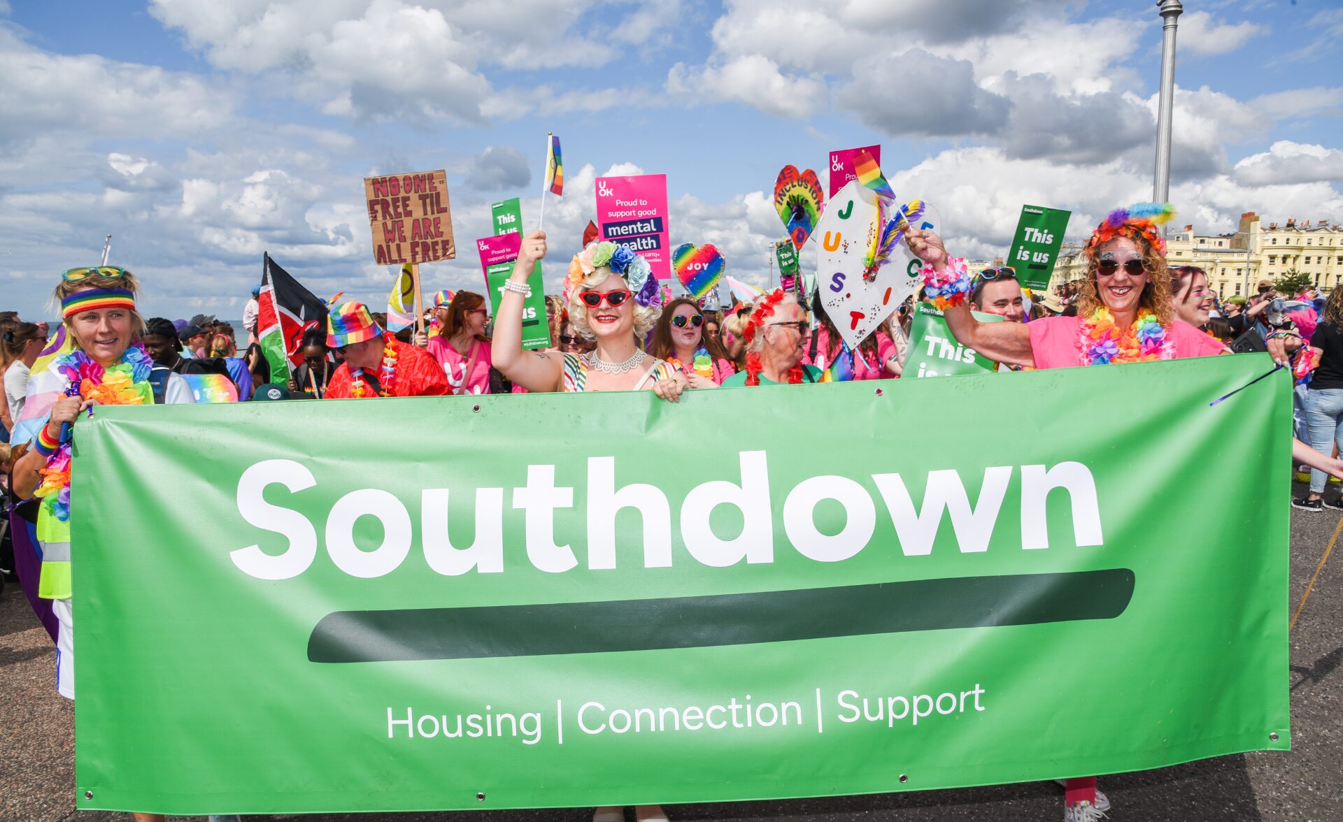 Southdown team marching in Brighton Pride Parade, holding a green Southdown banner with the words ‘Housing | Connection | Support’, surrounded by colourful rainbow outfits, flags, and inclusion signs.