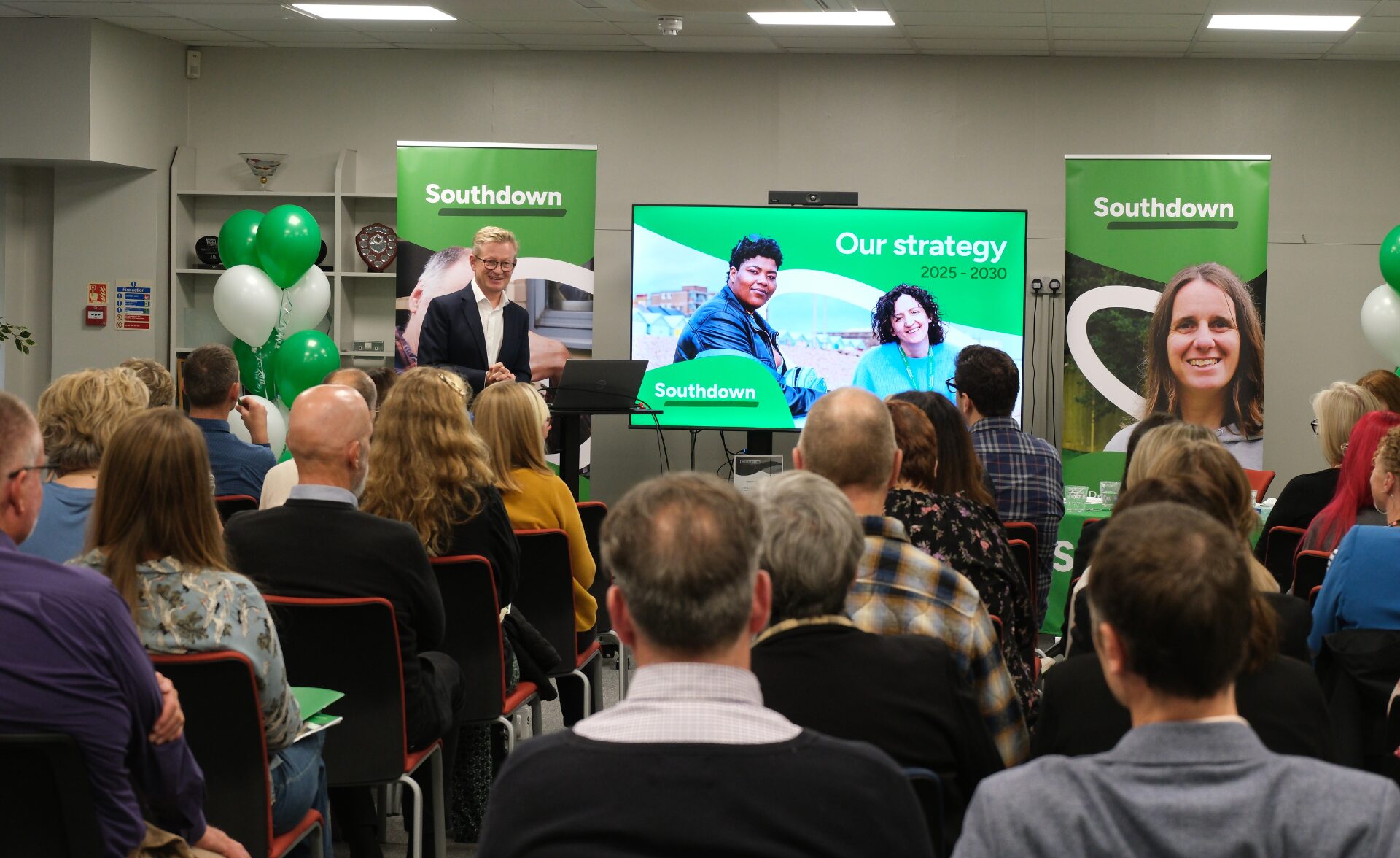 Southdown Chief Executive presenting the new Strategy 2025–2030 at the launch event in Lewes, with colleagues, clients and partners in attendance. A large screen displays the strategy branding alongside Southdown banners and balloons.