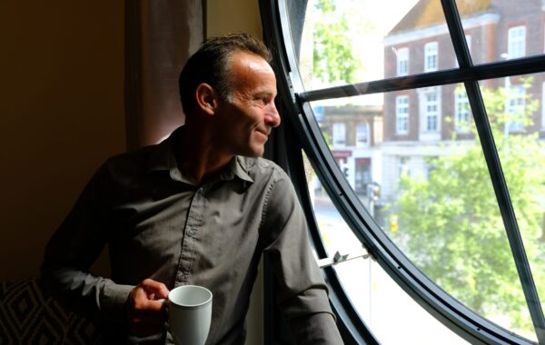 A man wearing a grey shirt holds a white mug and looks out of a large round window, softly lit by daylight. He smiles thoughtfully as he gazes outside at trees and buildings, reflecting a sense of hope and calm after finding support through Southdown Steps.