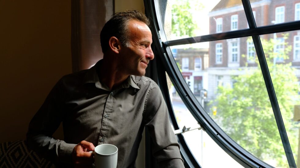 A man wearing a grey shirt holds a white mug and looks out of a large round window, softly lit by daylight. He smiles thoughtfully as he gazes outside at trees and buildings, reflecting a sense of hope and calm after finding support through Southdown Steps.