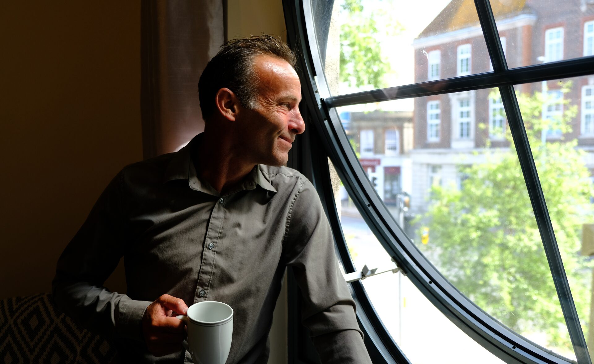 A man wearing a grey shirt holds a white mug and looks out of a large round window, softly lit by daylight. He smiles thoughtfully as he gazes outside at trees and buildings, reflecting a sense of hope and calm after finding support through Southdown Steps.