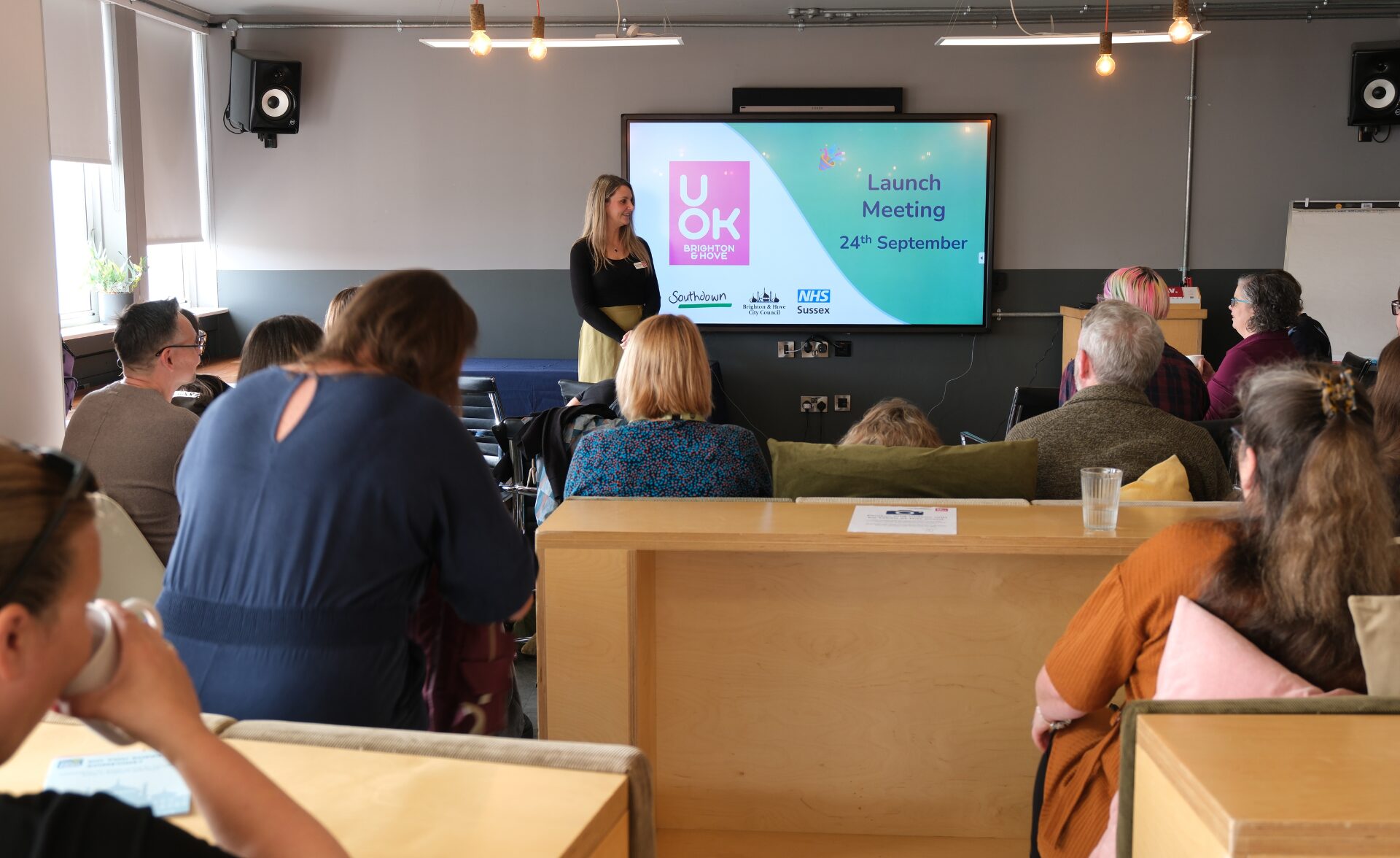 People attending the launch meeting for the UOK Brighton & Hove contract on 24th September, with a presentation screen displaying the UOK logo and partner organisations Southdown, Brighton & Hove City Council, and NHS Sussex.