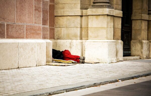 A person sleeping rough on the street, lying against the wall of a stone building — a representative image highlighting homelessness.