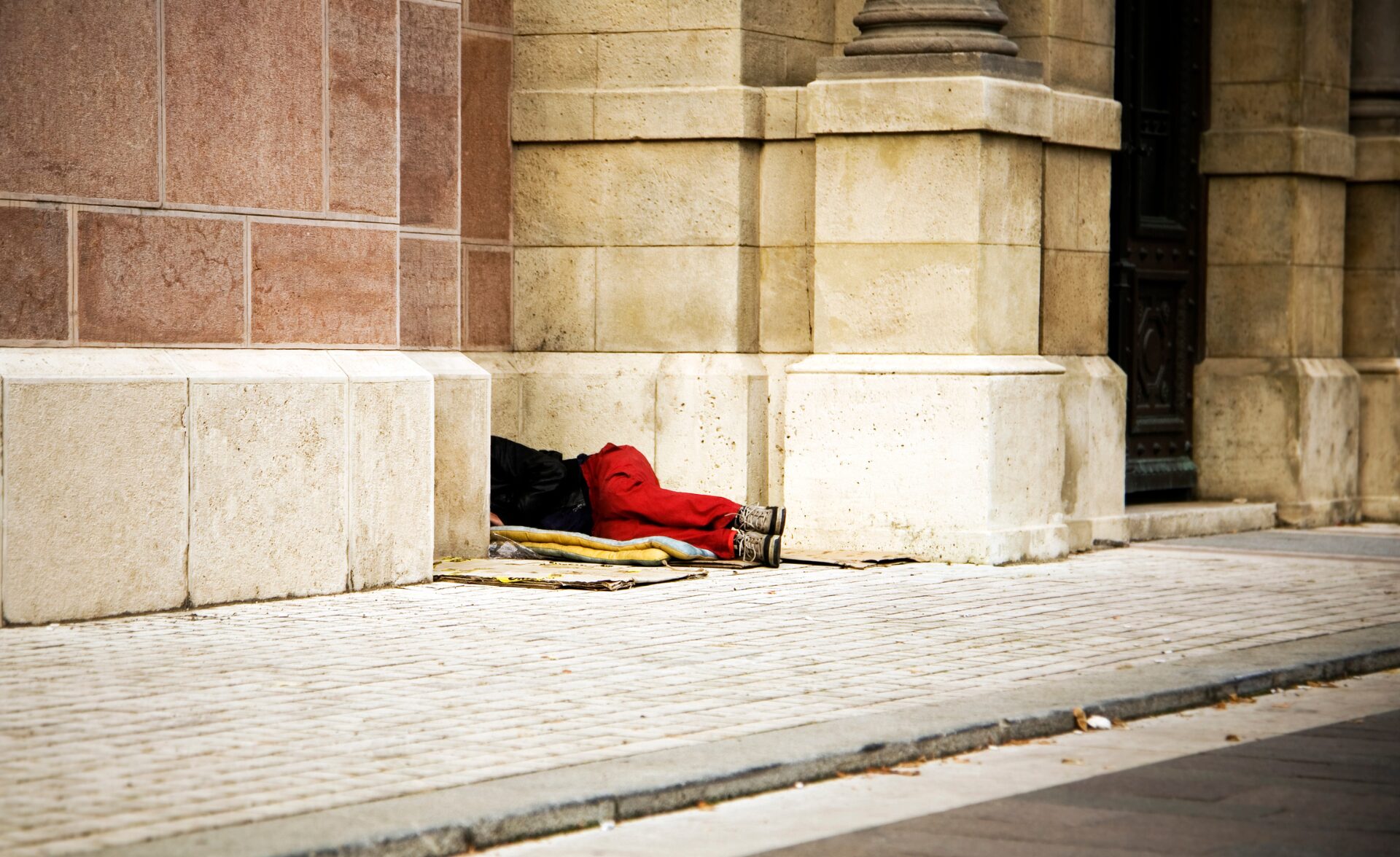 A person sleeping rough on the street, lying against the wall of a stone building — a representative image highlighting homelessness.