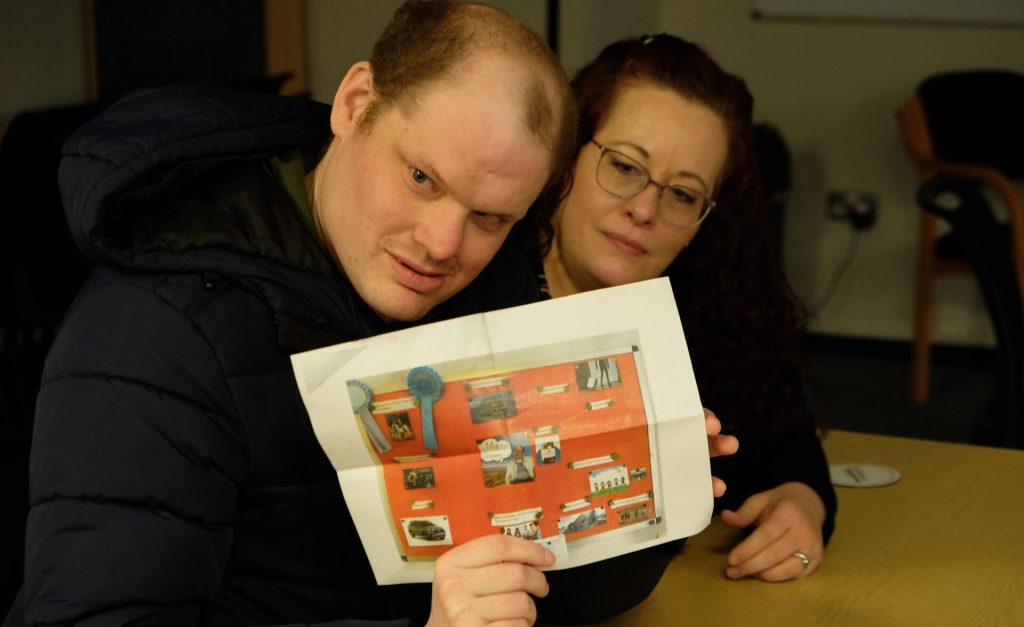 A man in a navy padded jacket sits at a table holding up a colourful poster with photos and blue rosettes. He leans slightly forward and looks directly at the camera. Next to him, a woman with long curly hair, glasses, and a black-and-white striped top sits closely, looking at the poster with a calm, supportive expression. The image is from a Southdown client involvement session.