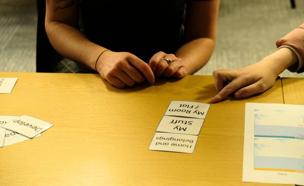 Two people seated at a table during a client involvement session, using visual cards with phrases like “My Room / Flat,” “My Stuff,” and “Home and Belongings” to support communication and explore topics related to quality of life for the redesigned MyPlan.