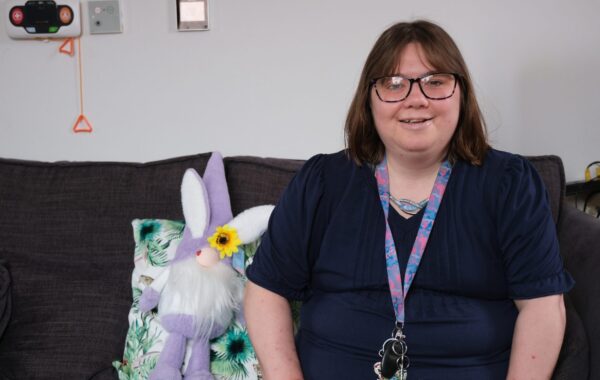 A woman with glasses sits on a sofa in a relaxed setting, smiling at the camera. Beside her is a decorative soft toy and patterned cushion. She is sharing her experience of receiving positive behaviour support and creating her own personalised support plan within a learning disability service.