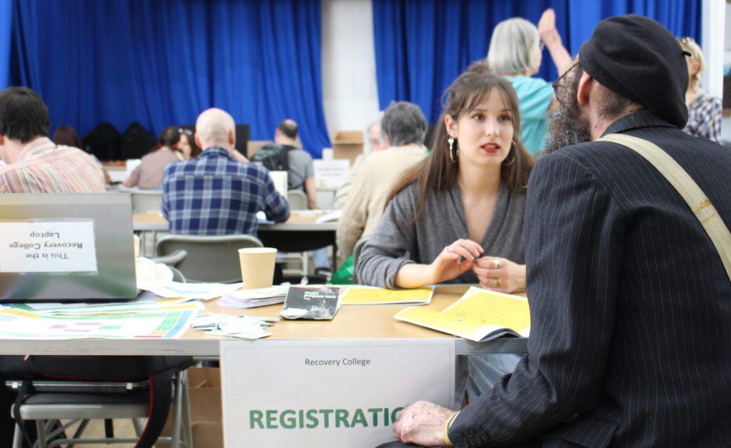 A Recovery College staff member speaks with a visitor at the registration desk during a busy Open Day event, with people chatting and exploring course materials in a welcoming community hall setting.