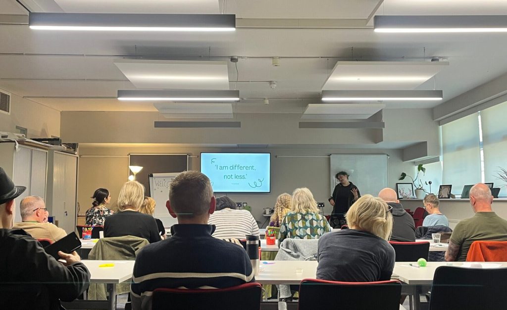 Professionals attend an Autism Awareness Workshop at the Recovery College, seated in a classroom while a presenter discusses autistic experiences. A screen at the front displays the quote 'I am different, not less,' highlighting the session's focus on understanding and accepting neurodiversity.