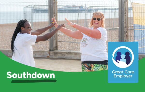 Two Southdown colleagues giving hi-five to each other while playing volleyball on a beach. Southdown logo and Great Care Employer icon sit on the foreground.