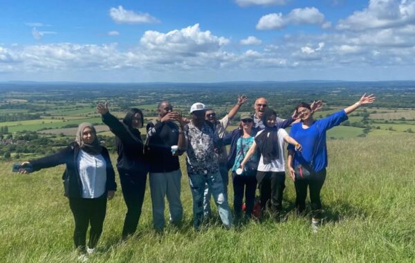 A group of people standing together on a grassy hill, smiling and waving, with a wide countryside view and blue sky behind them.