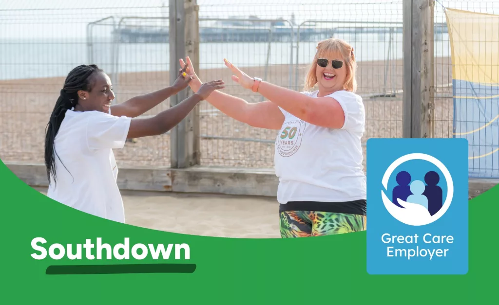 Two Southdown colleagues giving hi-five to each other while playing volleyball on a beach. Southdown logo and Great Care Employer icon sit on the foreground.