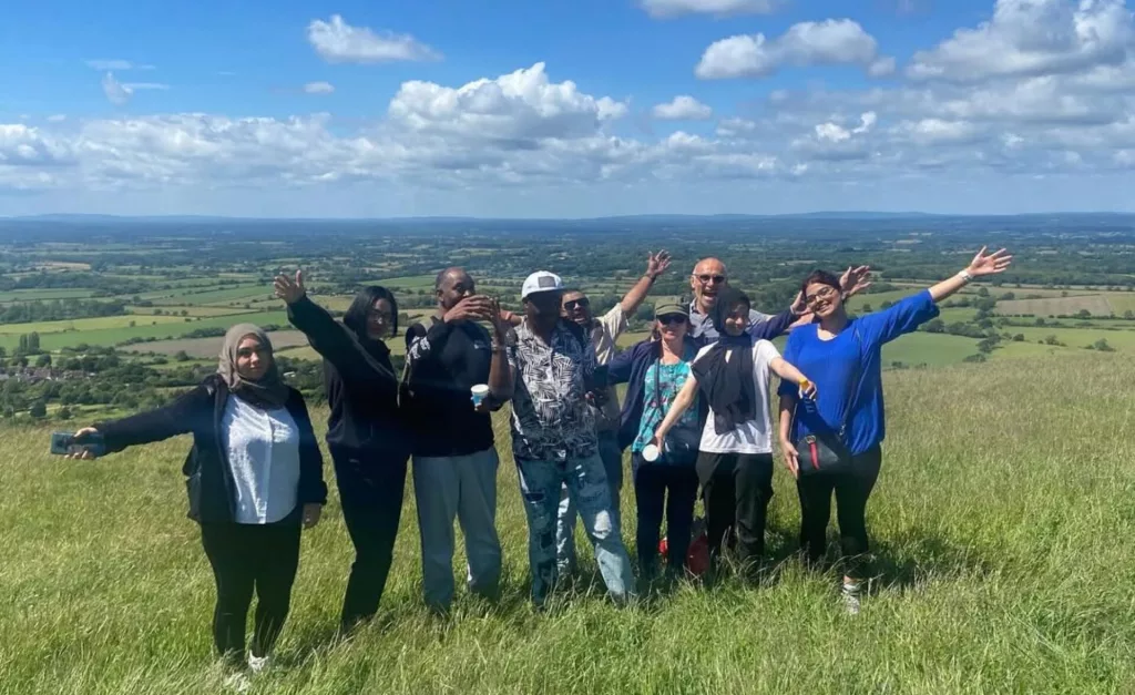 A group of people standing together on a grassy hill, smiling and waving, with a wide countryside view and blue sky behind them.